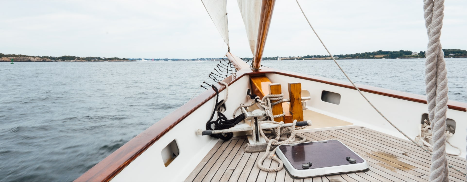 View from the bow of a sailing boat moving on water near Newport RI hotels, with visible ropes, wooden deck, and coastline in the distance.