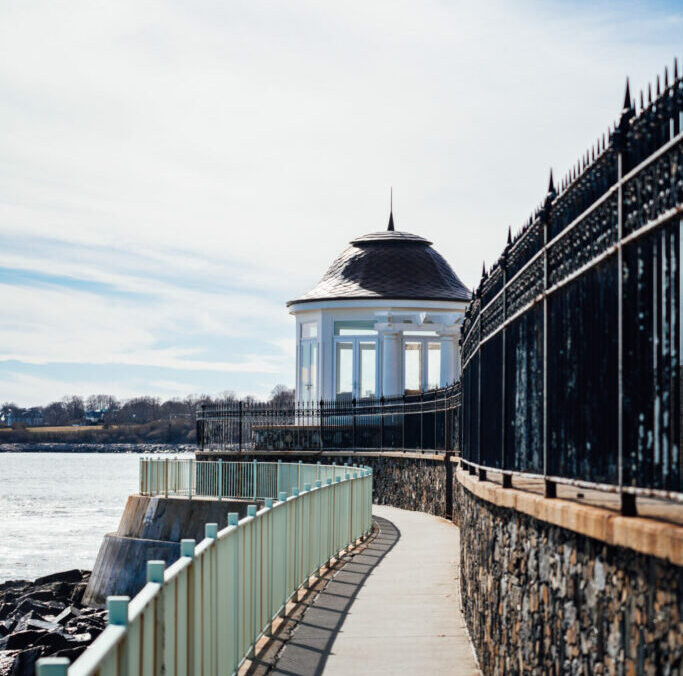 A coastal pathway in Newport, leading to a white gazebo, flanked by a black iron fence on one side and a green railing on the other, with calm water and clear skies in the background