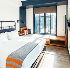 Modern bedroom in a Newport RI hotel with a large bed, gray and orange accents, hardwood floors, and large windows showing a city view.
