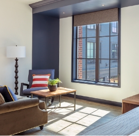 Modern living room with large window, blue and dark walls, a brown sofa with a striped pillow, a wooden coffee table, and a lamp in a Newport RI hotel.