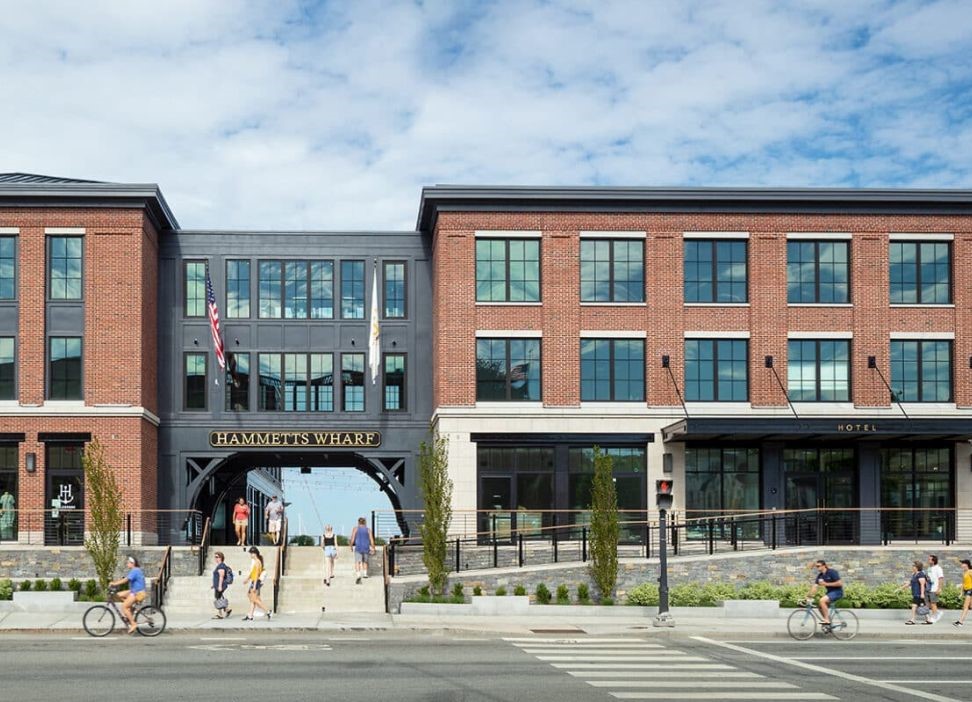 Modern brick building with large windows and "Hammet's Wharf Hotel" sign, featuring pedestrians and cyclists on a sunny day, is one of the notable hotels in Newport RI.