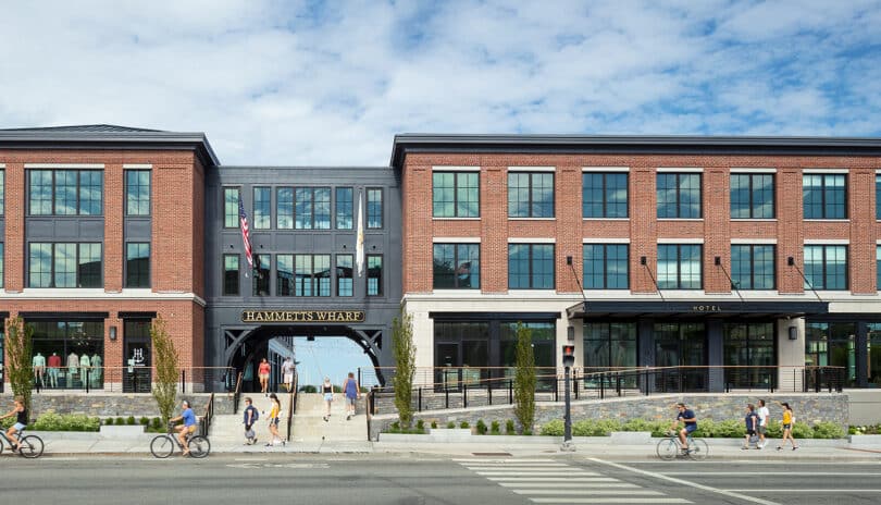 Modern two-story brick commercial building with large windows and a sign reading "hotels in Newport RI," pedestrians and cyclists passing in front.