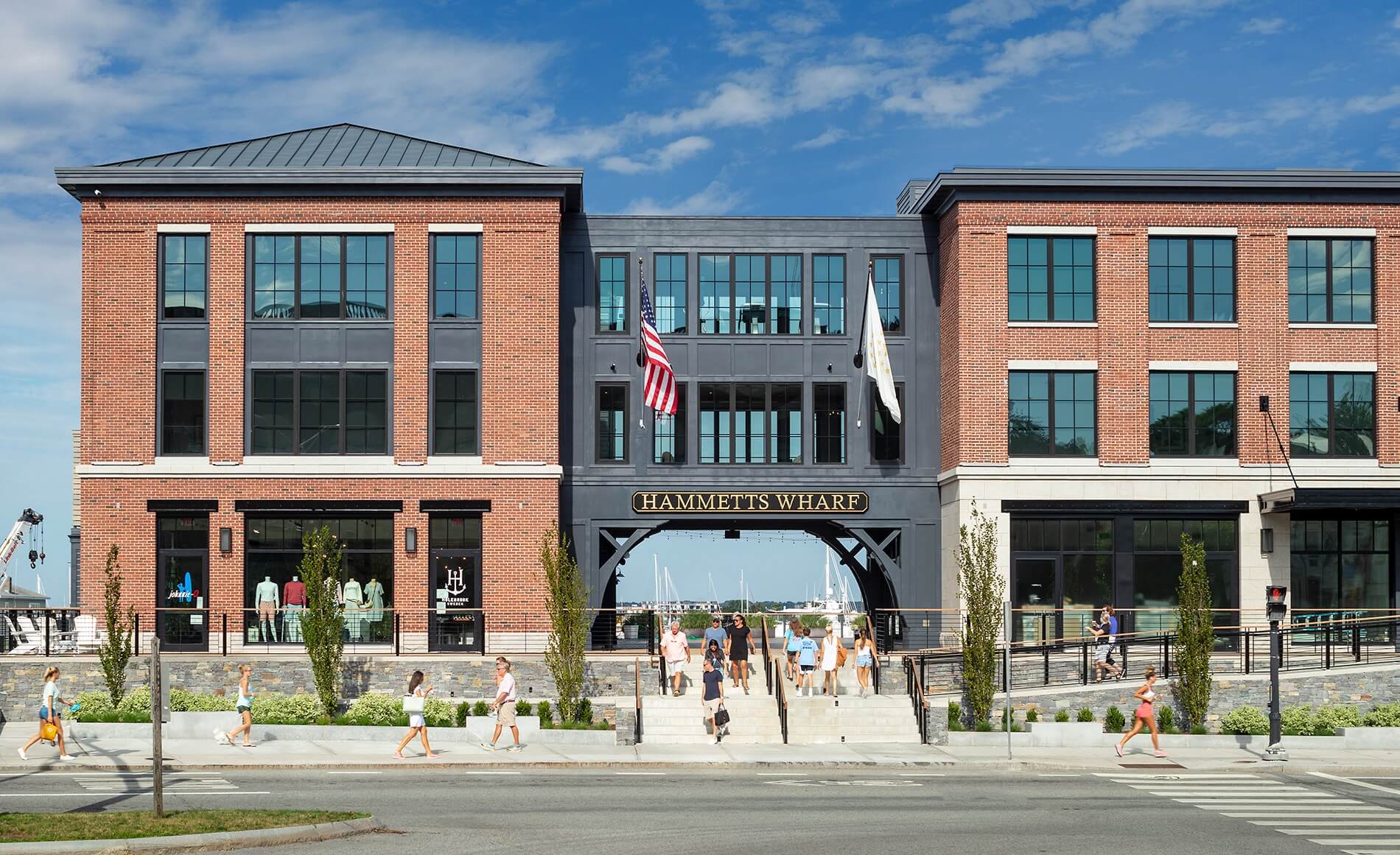 Modern two-story brick building with large glass windows, featuring an American flag and a sign that reads "Hammer's Wharf Hotel," with people walking in front. Located in Newport RI, this is
