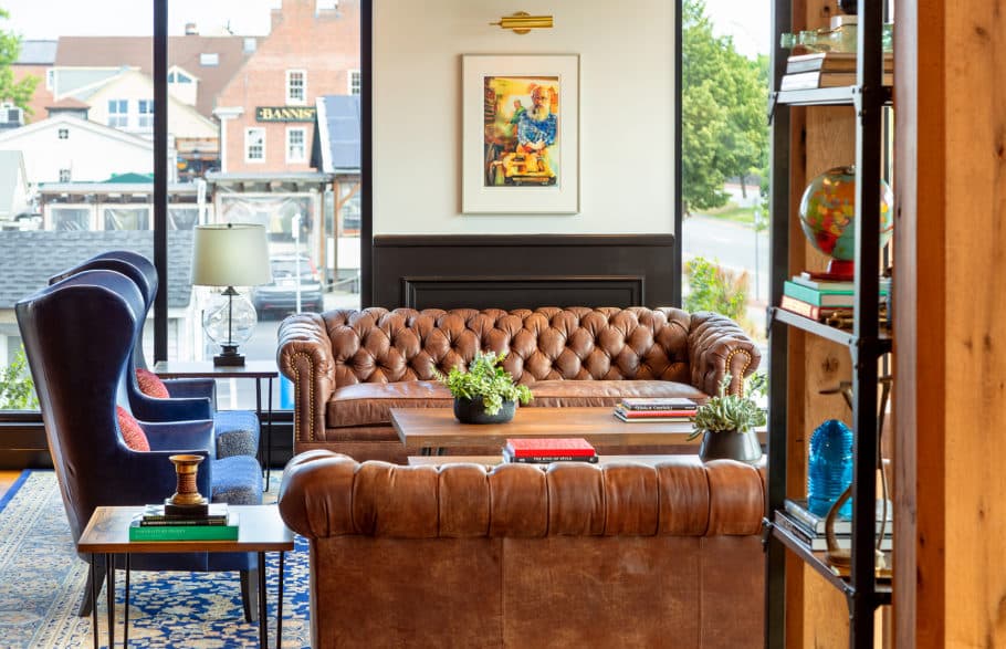 A cozy living room in a Newport RI hotel with a brown leather chesterfield sofa, a blue armchair, a small table with books, and a painting above a fireplace.