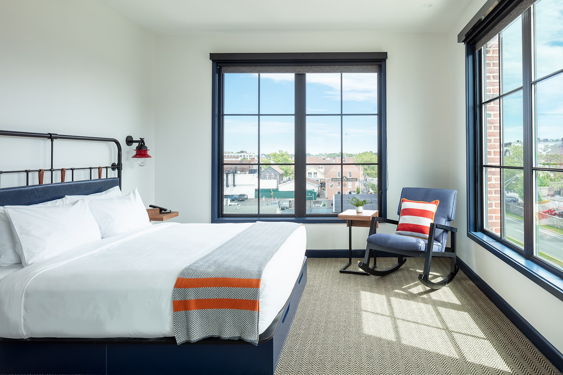 Modern hotel room in Newport RI featuring a large bed with striped blanket, a desk, a chair by the window, and a view of the cityscape outside.