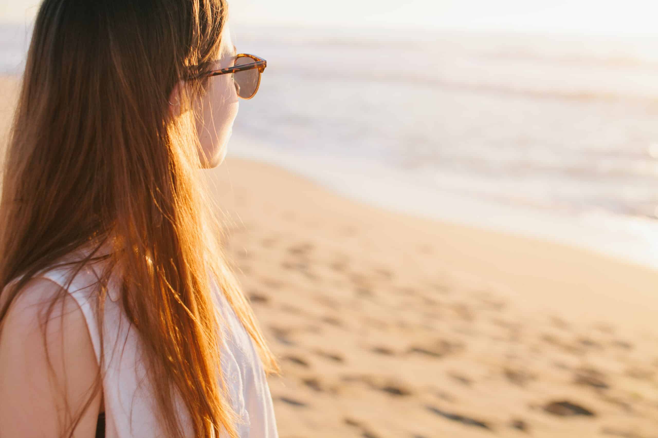 Woman with long hair and sunglasses looking out at the ocean from a sandy beach at sunset near a Newport RI hotel.
