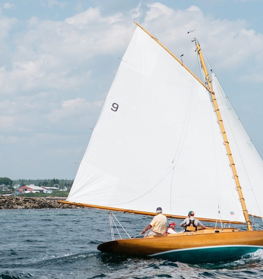 Two people sailing on a clear day in a wooden boat with a large white sail marked with the number 9, near a rocky shore close to the best hotels in Newport, RI.