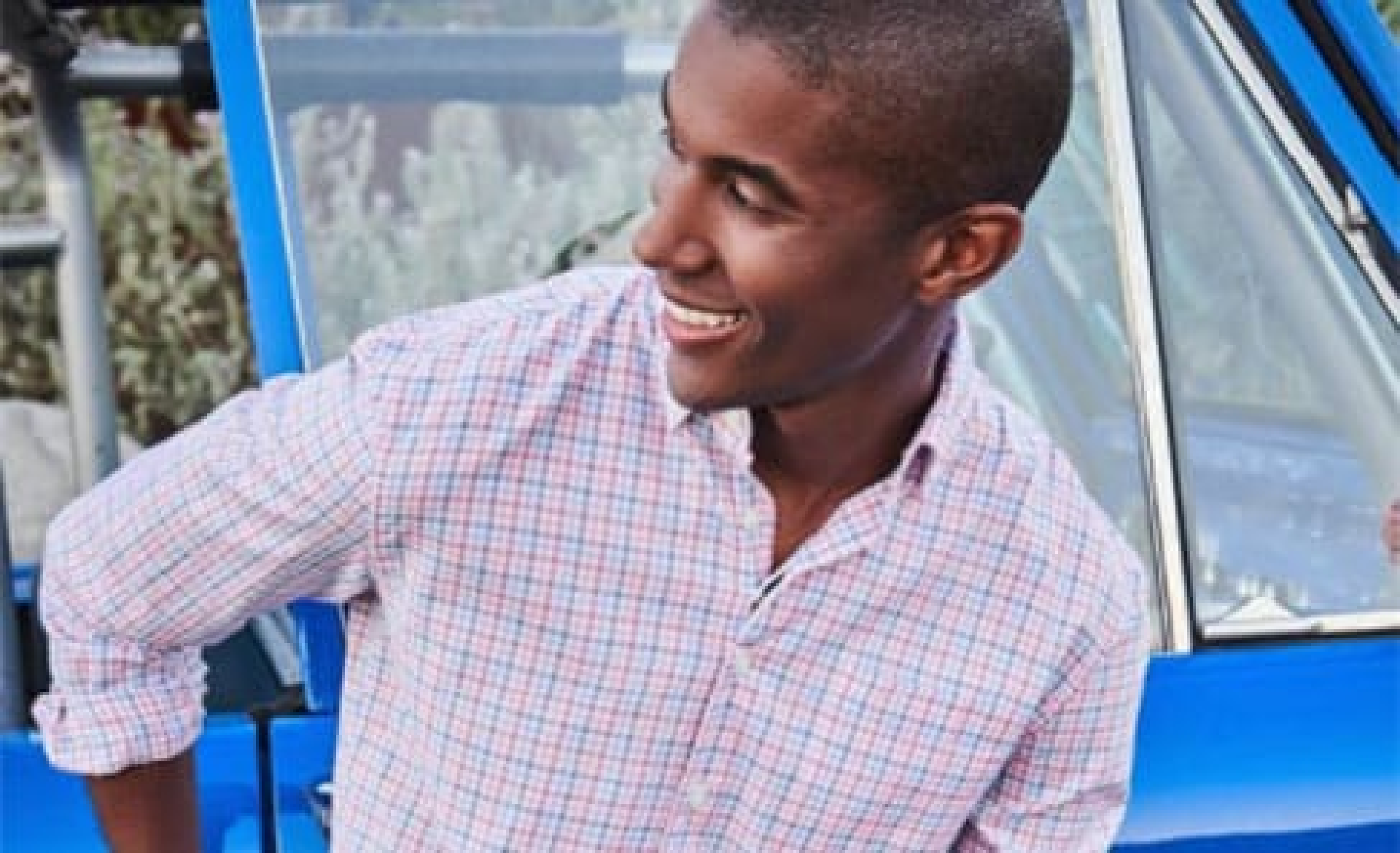 A smiling young man in a checkered shirt leaning on a blue car outside a Newport RI hotel.
