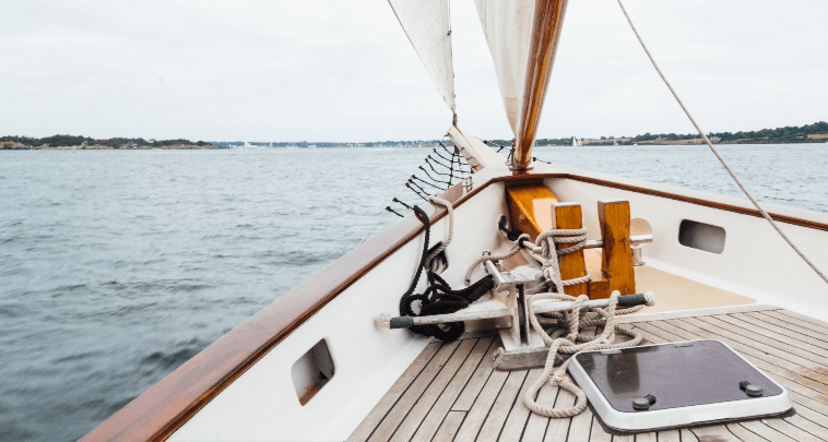 View from the bow of a sailing boat on a cloudy day, showing the deck, ropes, and open water with a distant shoreline near hotels in Newport RI.