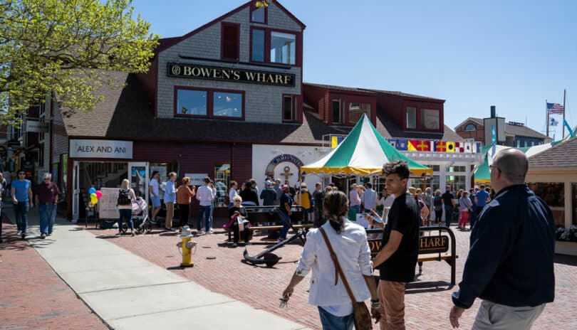 People walking and mingling near Bowen's Wharf, featuring a bustling street scene with shops, hotels in Newport RI, and a vendor tent under a clear sky.
