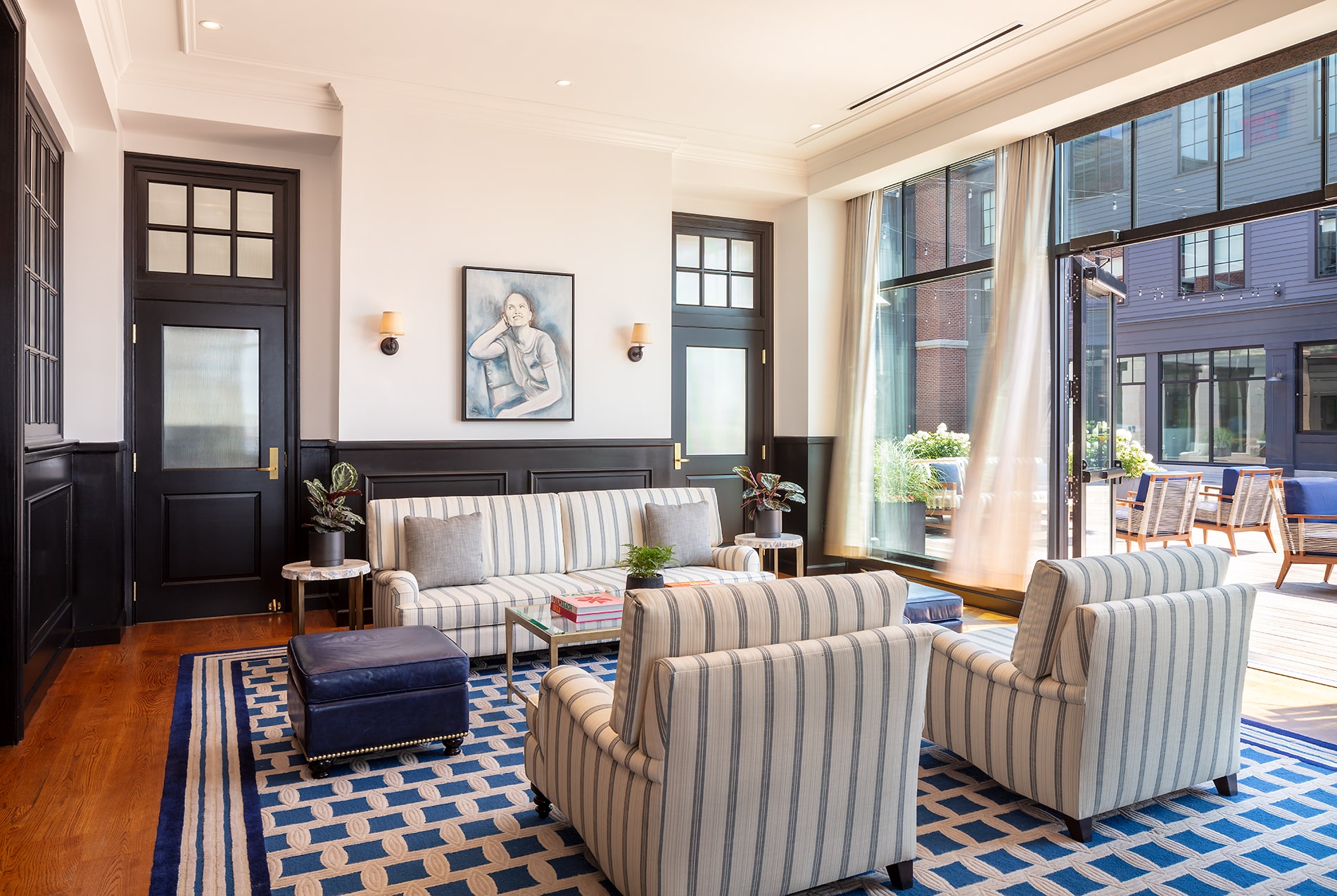 Modern living room with striped sofas, blue rug, and large windows leading to a balcony in a Newport RI hotel. A portrait hangs above a black console table.
