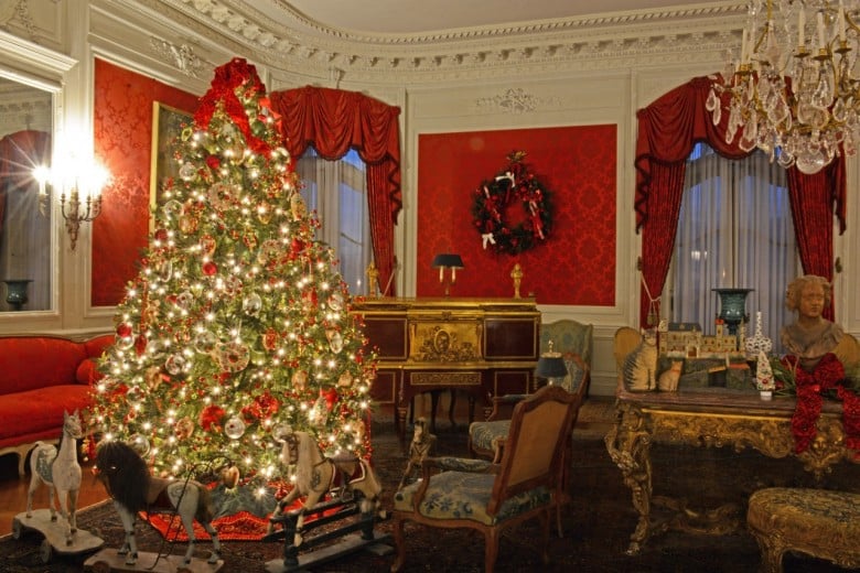 An elegantly decorated Christmas tree stands in a richly furnished room at a Newport RI hotel, with red walls, antique furniture, and a chandelier.