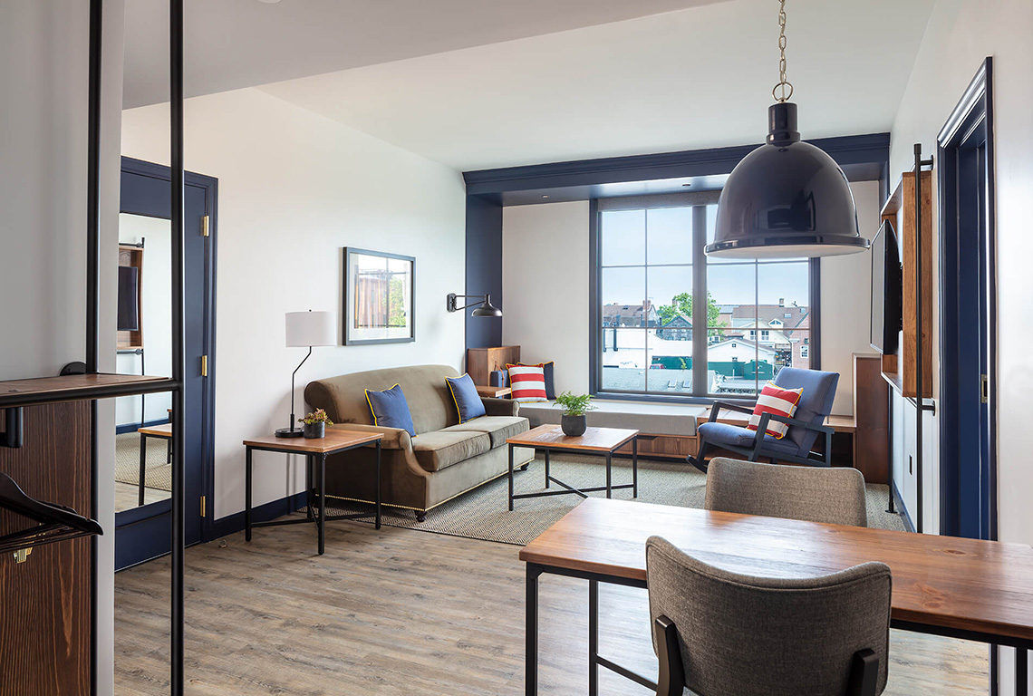 Modern living room with a tan sofa, two blue and red accent chairs, a large pendant light, and a dining area in the background of a Newport RI hotel.