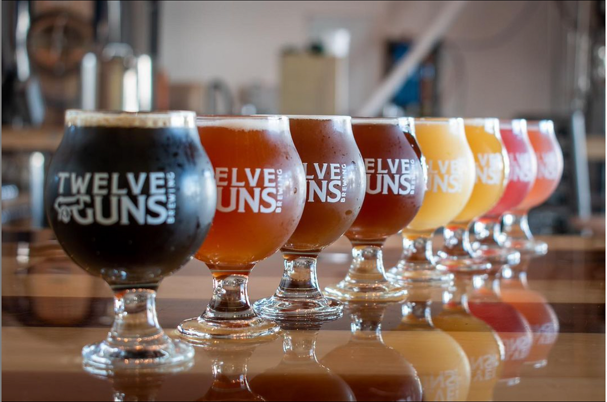 A row of various beers in different shaped glasses, labeled "twelve guns," arranged on a bar counter at a Newport RI hotel, showcasing a gradient from dark to light colors.