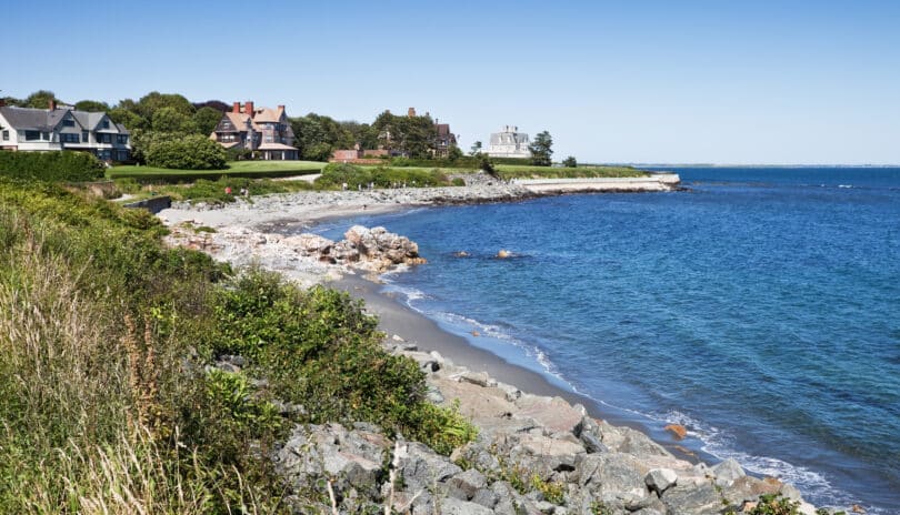 Coastal landscape showing a rocky beach with hotels in the background under a clear blue sky.