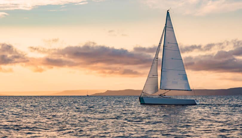 A sailboat sails on calm waters at sunset with a clear sky and distant hills on the horizon near Newport, RI.