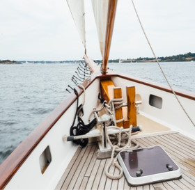 View from the bow of a sailboat with wooden deck and white sails, sailing near the coastline of Newport RI under a cloudy sky.