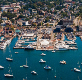 Aerial view of a coastal town with a marina filled with boats, surrounded by densely packed buildings and greenery, not far from popular hotels in Newport, RI.
