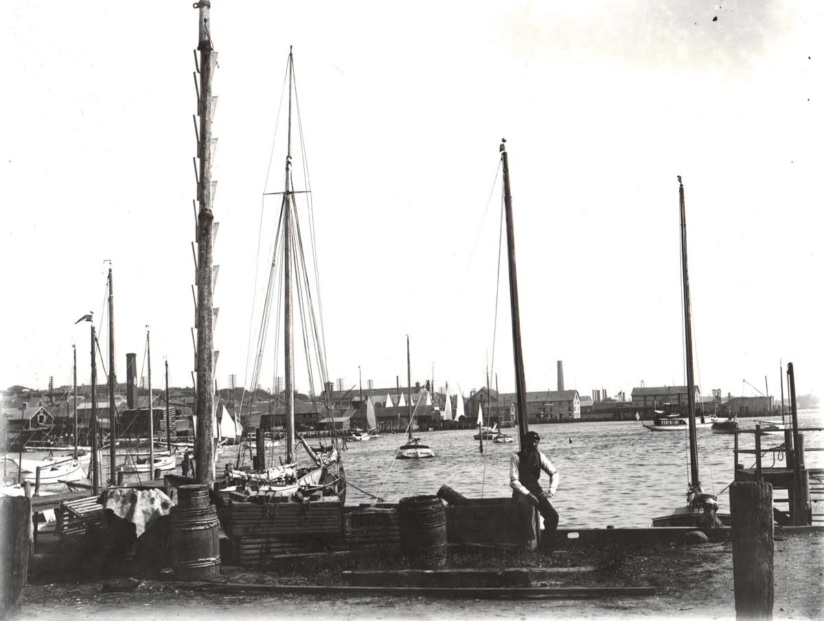 Black and white photo of a man walking near docked sailboats in a busy port area with hotels in Newport RI in the background.