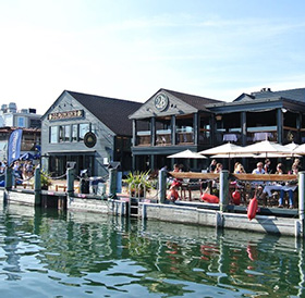 Waterfront dining scene with people sitting at tables in front of rustic buildings, under a clear blue sky, near popular hotels in Newport RI.