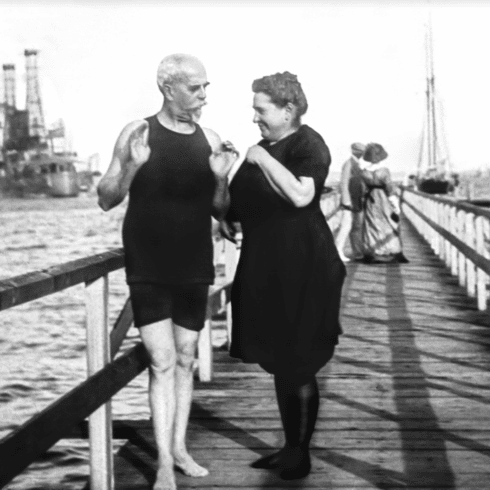 An elderly couple happily conversing on a pier in Newport, RI; the man in a swimsuit and the woman in a dress, with water and boats in the background.