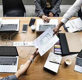 A bird's-eye view of a collaborative workspace in a Newport RI hotel, with four individuals discussing over a document, surrounded by laptops, books, and coffee cups.