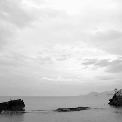 Black and white image of a calm sea with a sunken ship partially visible near the shore under a cloudy sky, close to hotels in Newport RI.