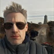 Man with gray hair wearing sunglasses in the foreground, a rural road and a tower near Newport RI hotels in the background.