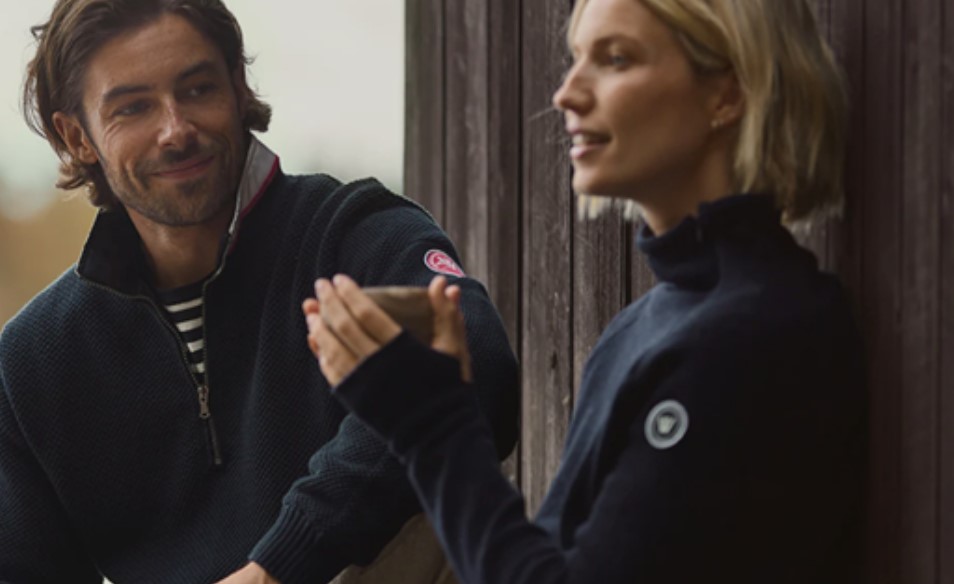 A man smiling at a woman who is clapping her hands, both dressed in casual jackets, standing by a wooden door of a hotel in Newport RI.