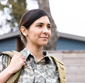 A woman in military uniform with a backpack, smiling slightly and looking off to the side, stands outdoors near a popular Newport RI hotel.