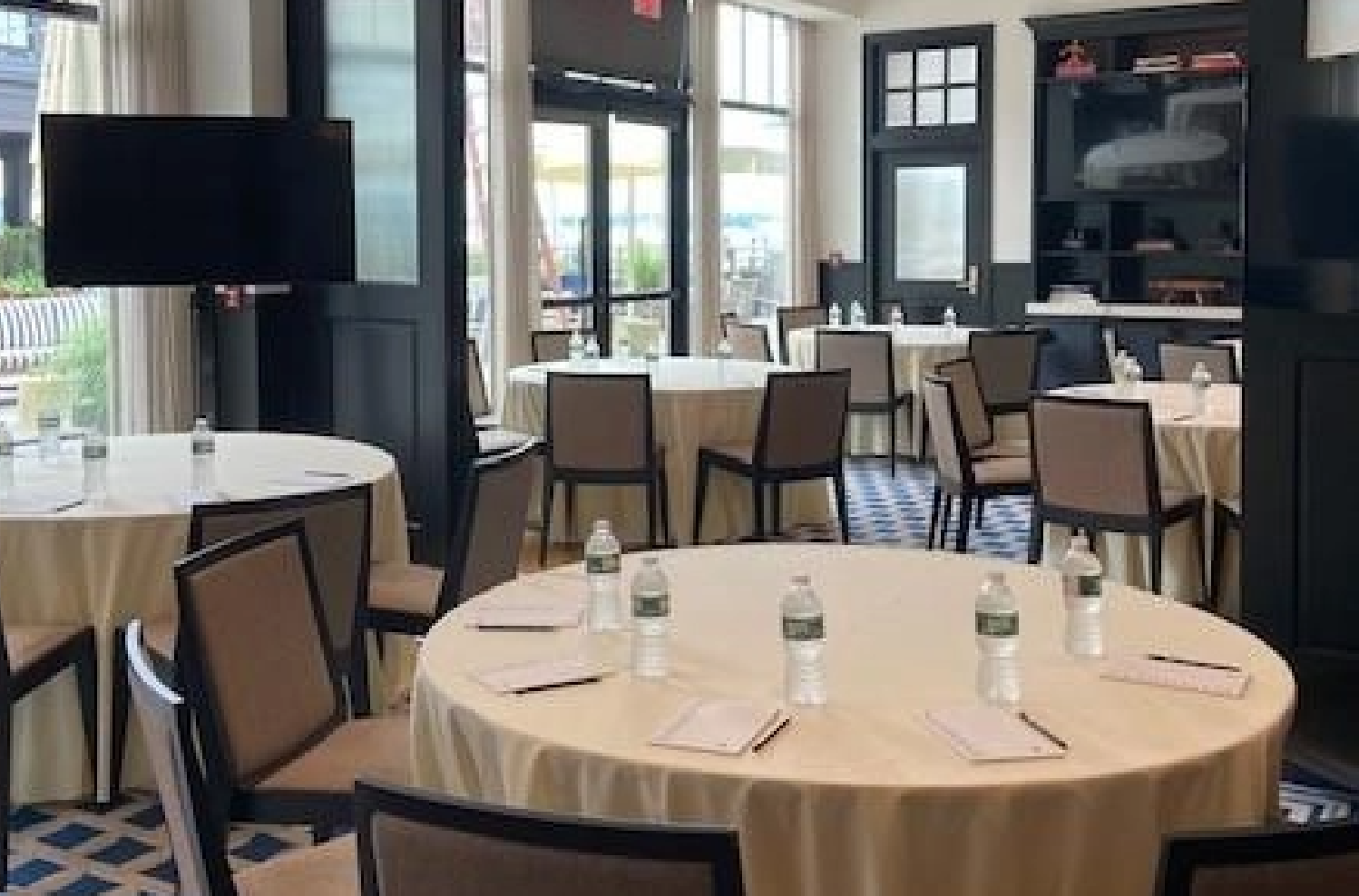 A conference room in a Newport, RI hotel with round tables set up for a meeting, featuring water bottles and notepads on each table, surrounded by chairs and large windows.