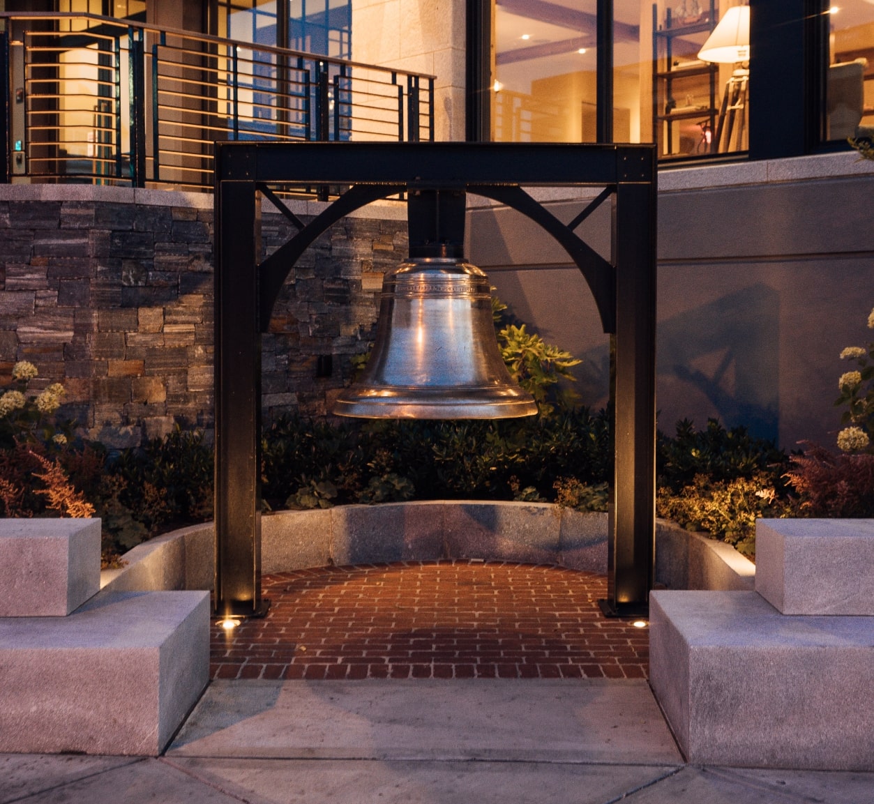 A large bell mounted at the center of a paved area with benches, illuminated by warm lights at dusk, near several places to stay in Newport.