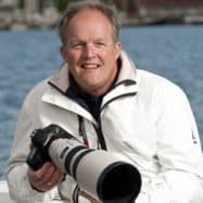 A smiling middle-aged man holding a camera with a long lens, wearing a white jacket, sitting by a body of water near a Newport RI hotel.