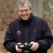 Man in glasses and a black jacket holding a drone controller in a forested area near Newport RI hotels.