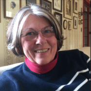A smiling woman wearing glasses and a black and white striped turtleneck, sitting indoors with framed pictures in the background at a hotel in Newport RI.