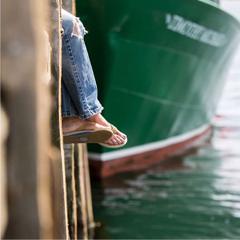 Person sitting on a dock in Newport, RI with their feet dangling above the water, beside a green and red boat.