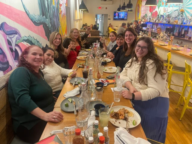 A group of women sitting at a long table in a restaurant, smiling and enjoying their meal.